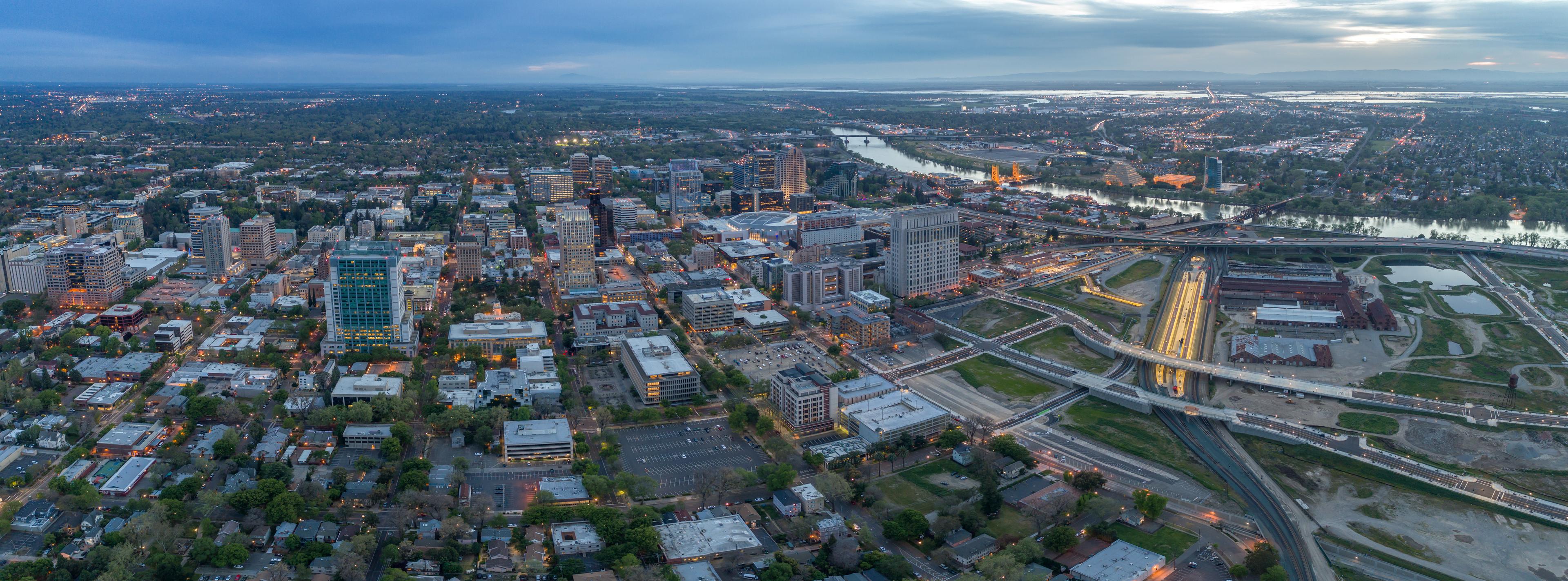 An expansive aerial view of downtown Sacramento, California, during twilight. The city's skyline is illuminated with buildings of various heights, and the Tower Bridge is lit up in the background, spanning the Sacramento River. Roads and highways crisscross the landscape, leading into surrounding neighborhoods and green spaces.