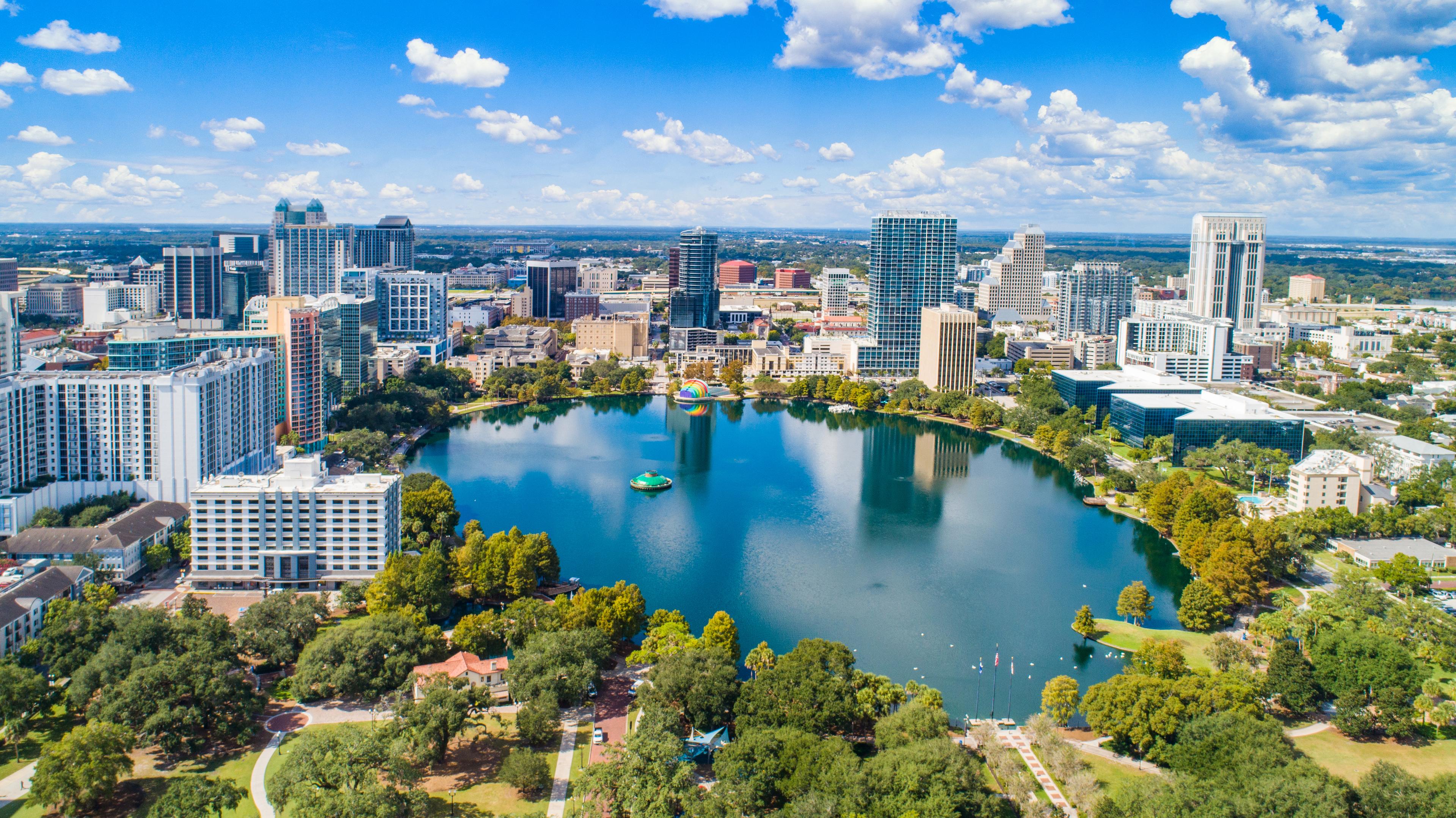 Aerial view of Lake Eola Park in downtown Orlando, surrounded by high-rise buildings and lush greenery under a bright blue sky.