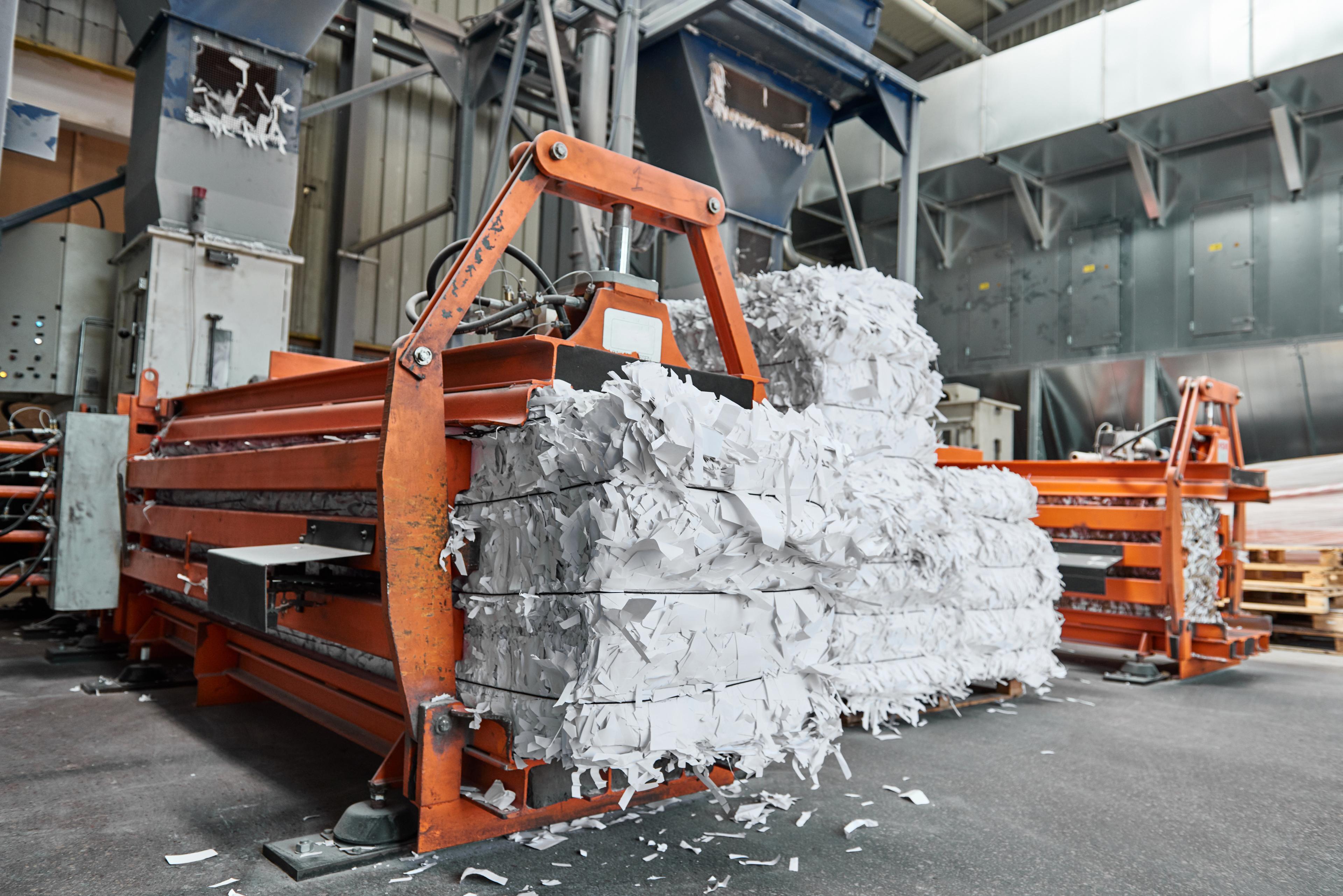 Industrial paper shredding machine processing and compacting shredded paper into large bales in a factory setting. The machine is surrounded by stacks of compressed shredded paper, indicating a high-volume shredding and recycling operation.