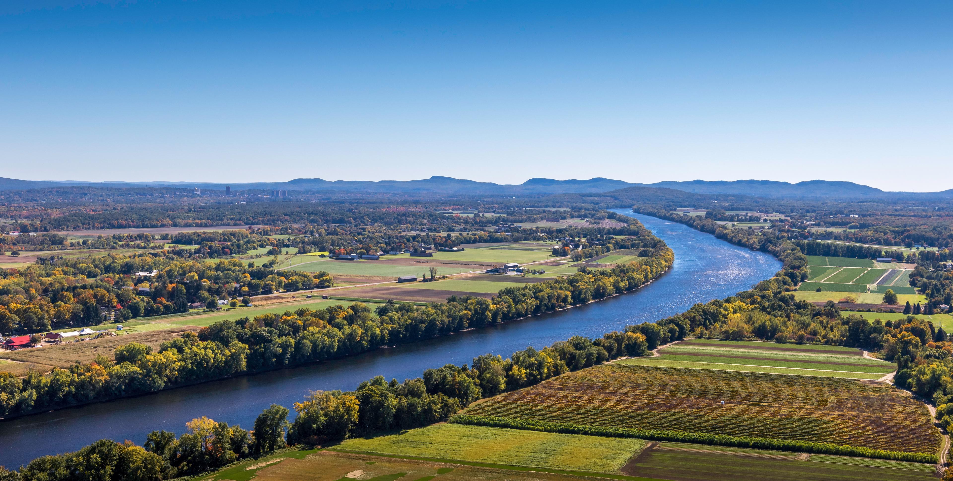 An aerial view of the Connecticut River meandering through lush farmland and vibrant forests, with distant hills and a clear blue sky in the background.