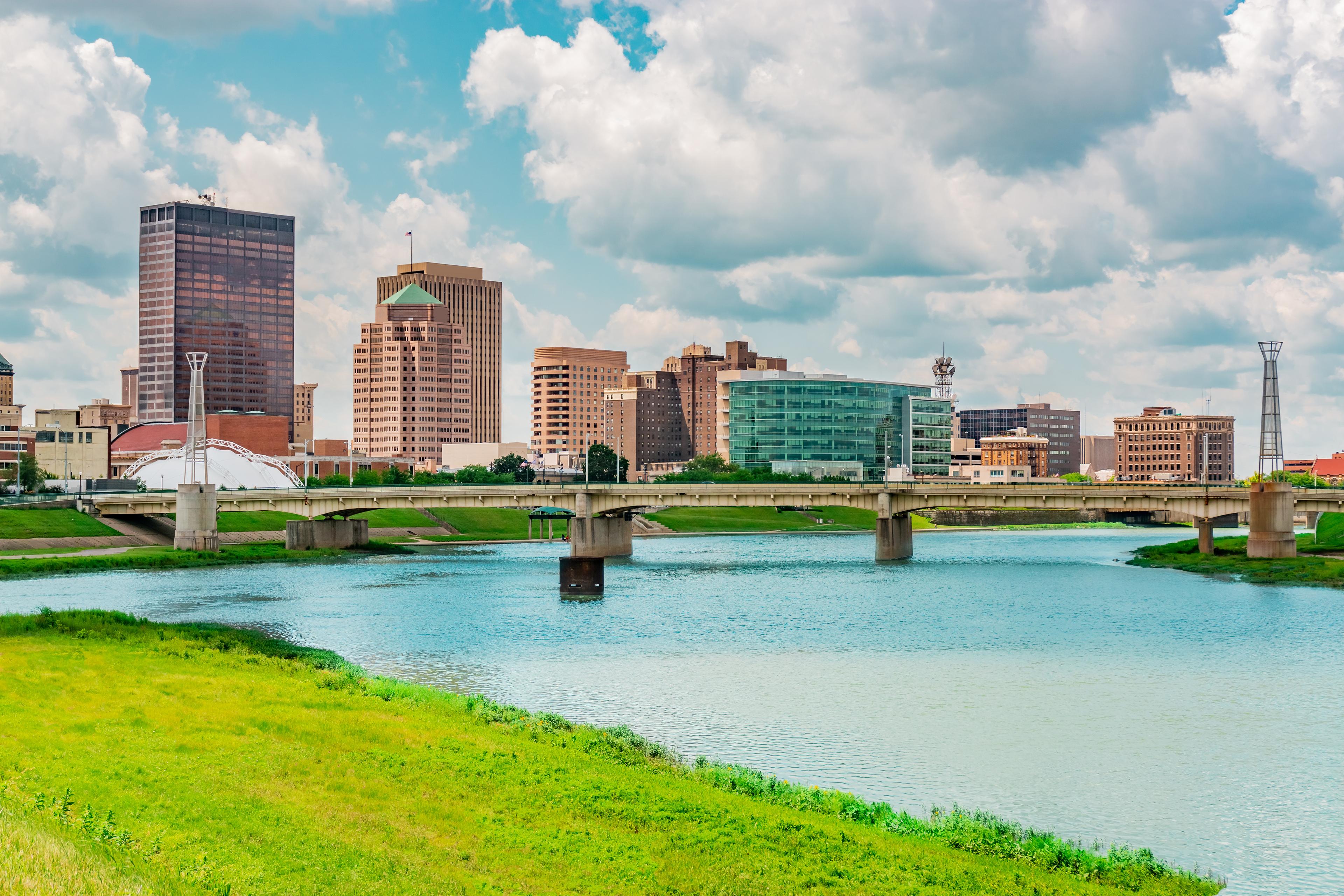 Daytime view of downtown Dayton, Ohio, featuring a bridge spanning the Great Miami River and a mix of modern and historic buildings under a partly cloudy sky.