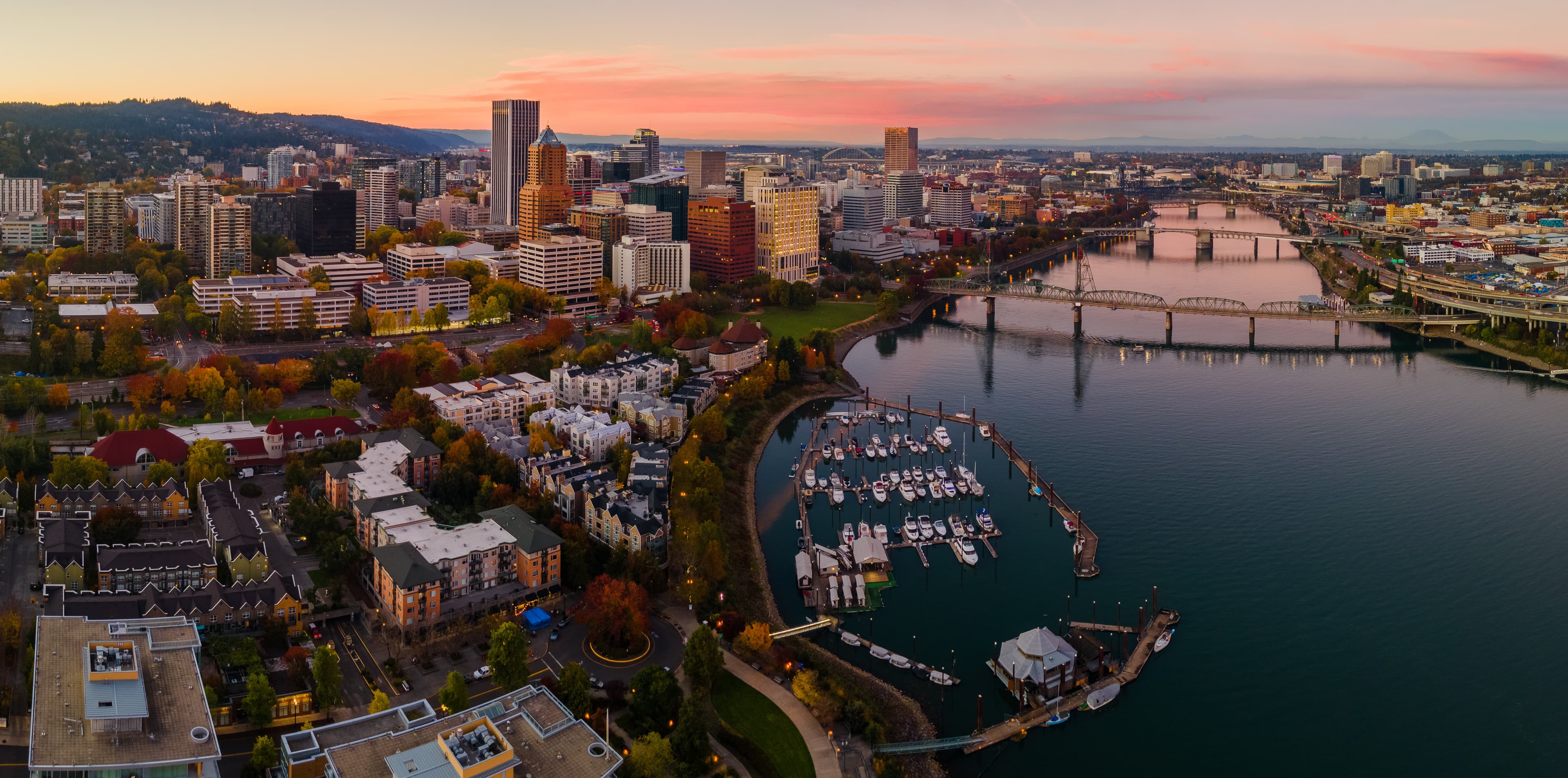 Downtown Portland, Oregon at sunset, featuring high-rise buildings, waterfront parks, and marina boats along the Willamette River with bridges crossing in the distance.