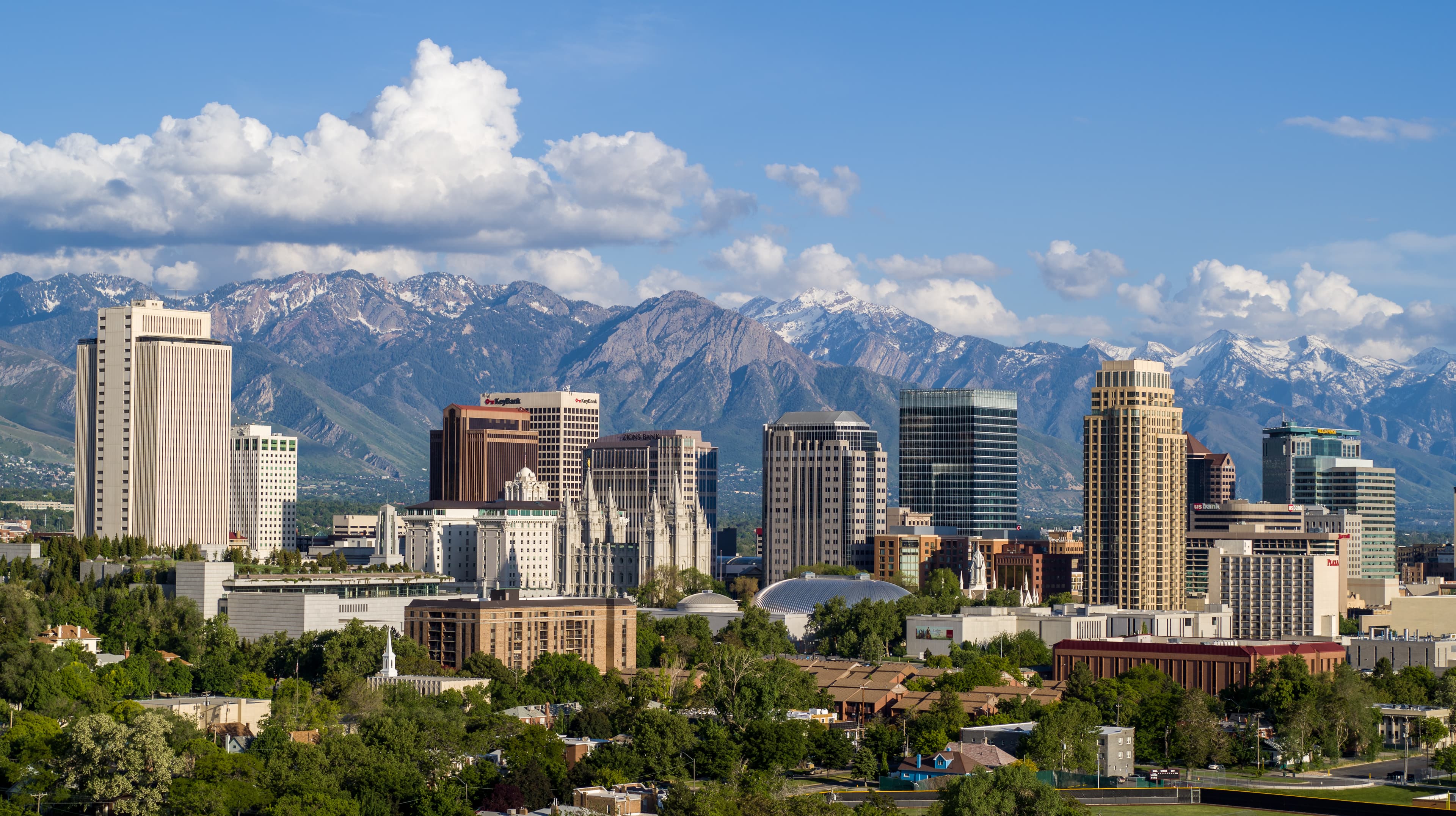Skyline of Salt Lake City, Utah, with modern high-rise buildings in the foreground and the snow-capped Wasatch Mountains towering in the background under a bright blue sky dotted with fluffy white clouds.