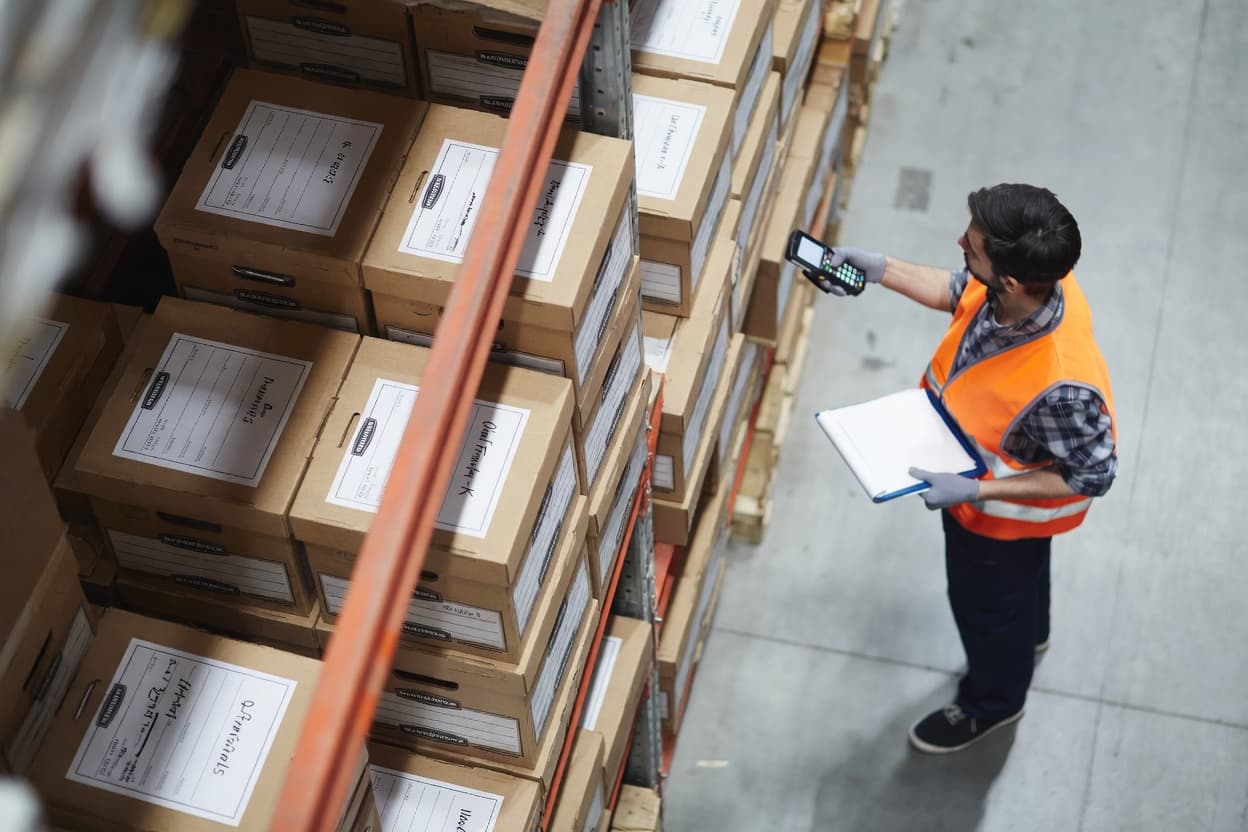Warehouse worker scanning labeled records boxes with a handheld barcode device, representing inventory tracking, records storage management, and real-time visibility in a records center.