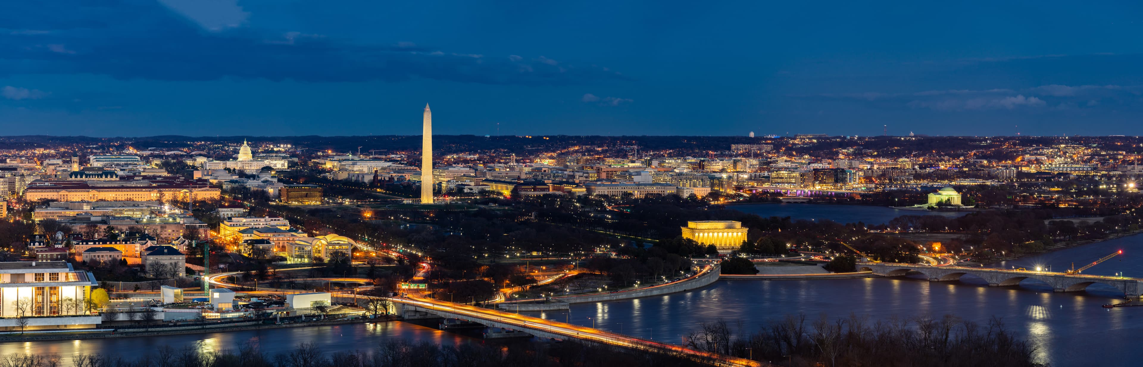 A panoramic nighttime view of Washington, D.C., featuring the illuminated Washington Monument, U.S. Capitol, Lincoln Memorial, Jefferson Memorial, and bridges crossing the Potomac River, with city lights glowing under a deep blue sky.