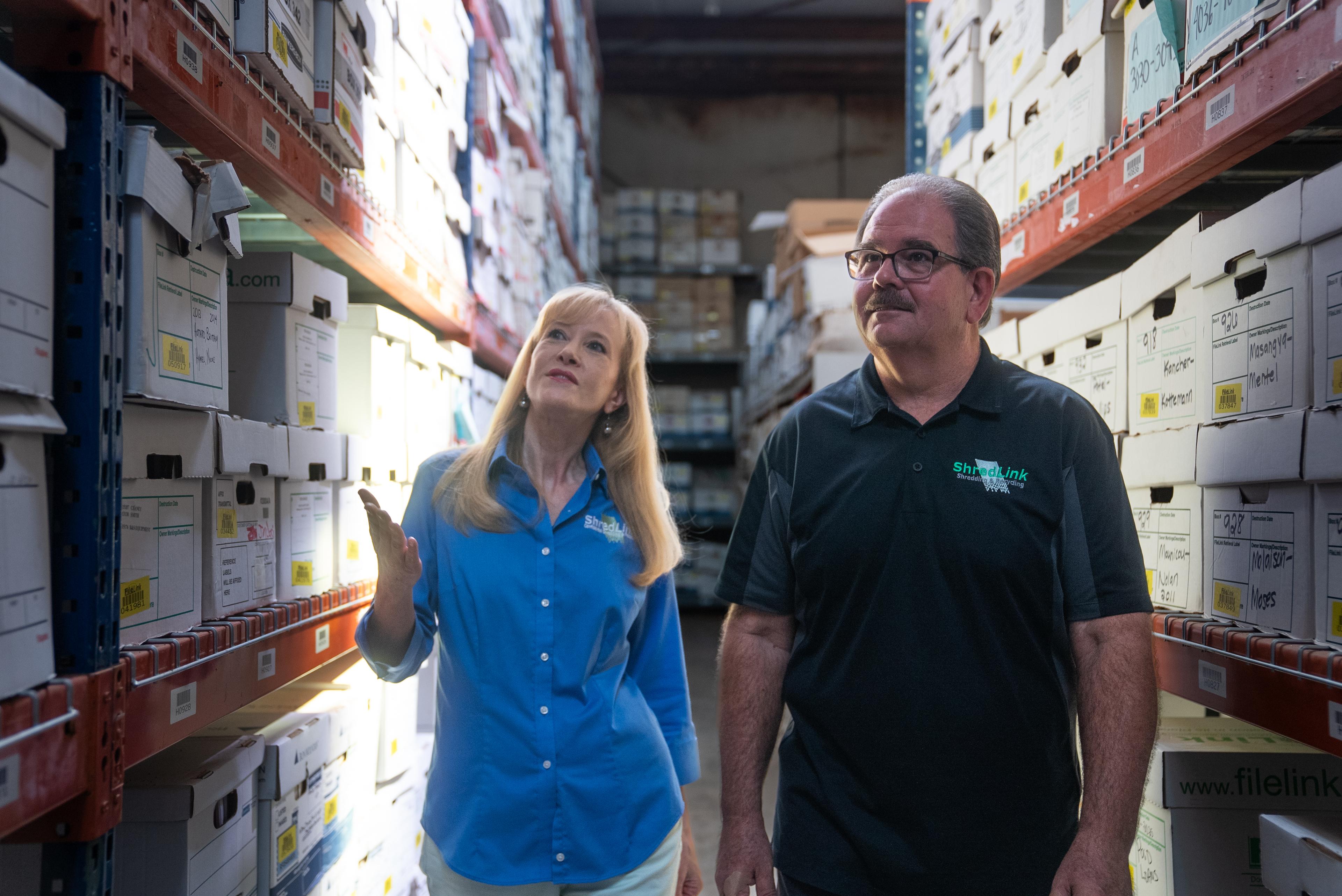Two professionals walking through a secure records storage facility, inspecting rows of labeled archive boxes for compliance and inventory management.