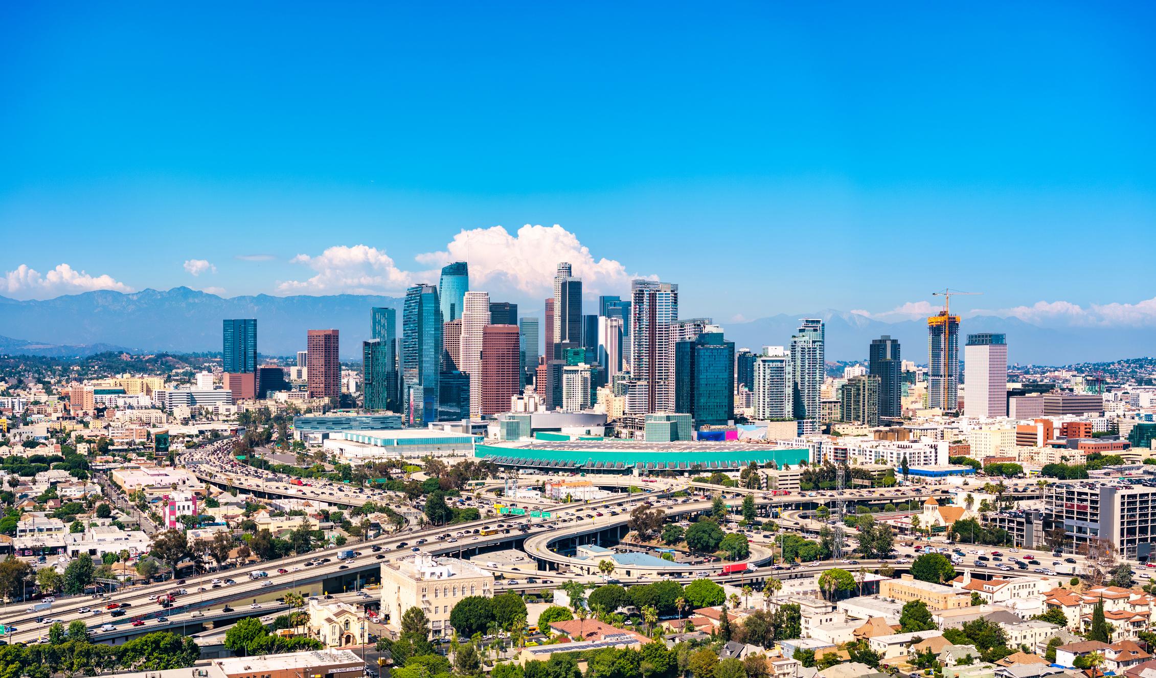 Panoramic view of downtown Los Angeles skyline with iconic high-rise buildings, bustling freeway system, and clear blue skies, showcasing Southern California urban infrastructure.