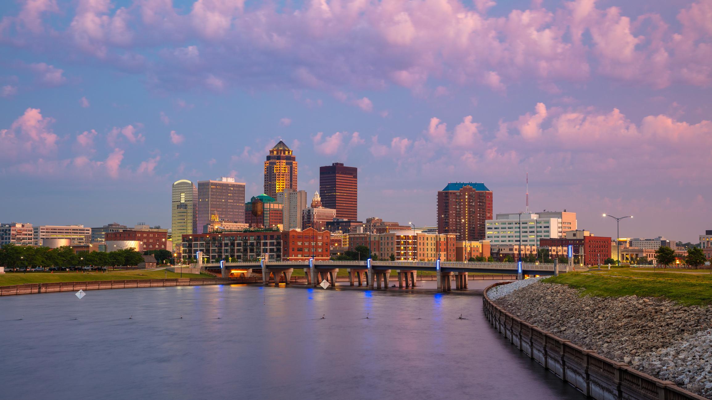 A view of the Des Moines, Iowa skyline at sunset, featuring tall buildings and vibrant colors reflected in the calm river. The scene includes a bridge and a grassy riverbank under a pink and purple cloud-filled sky.