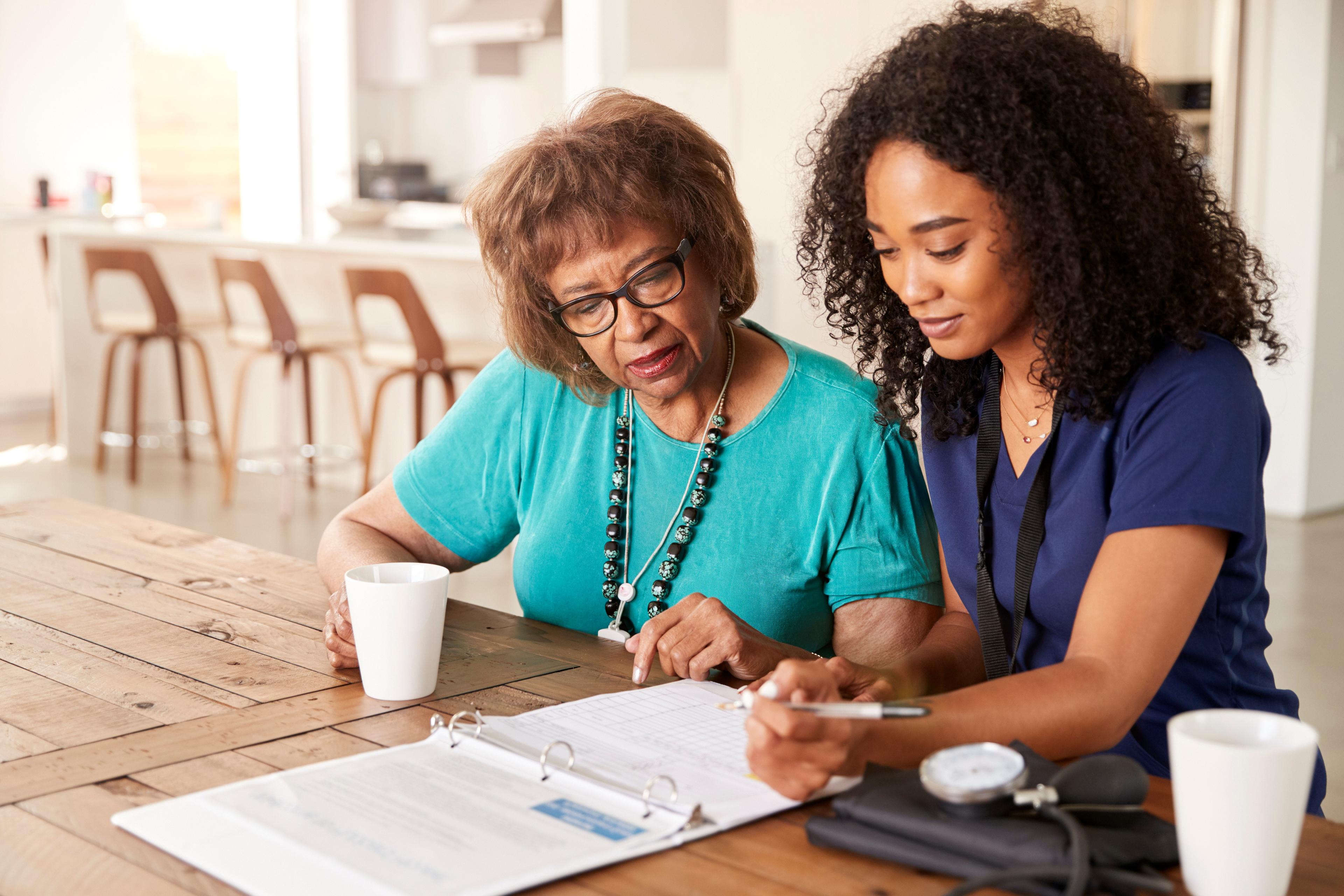 Younger healthcare worker assists an older woman with documents at a kitchen table, both holding mugs