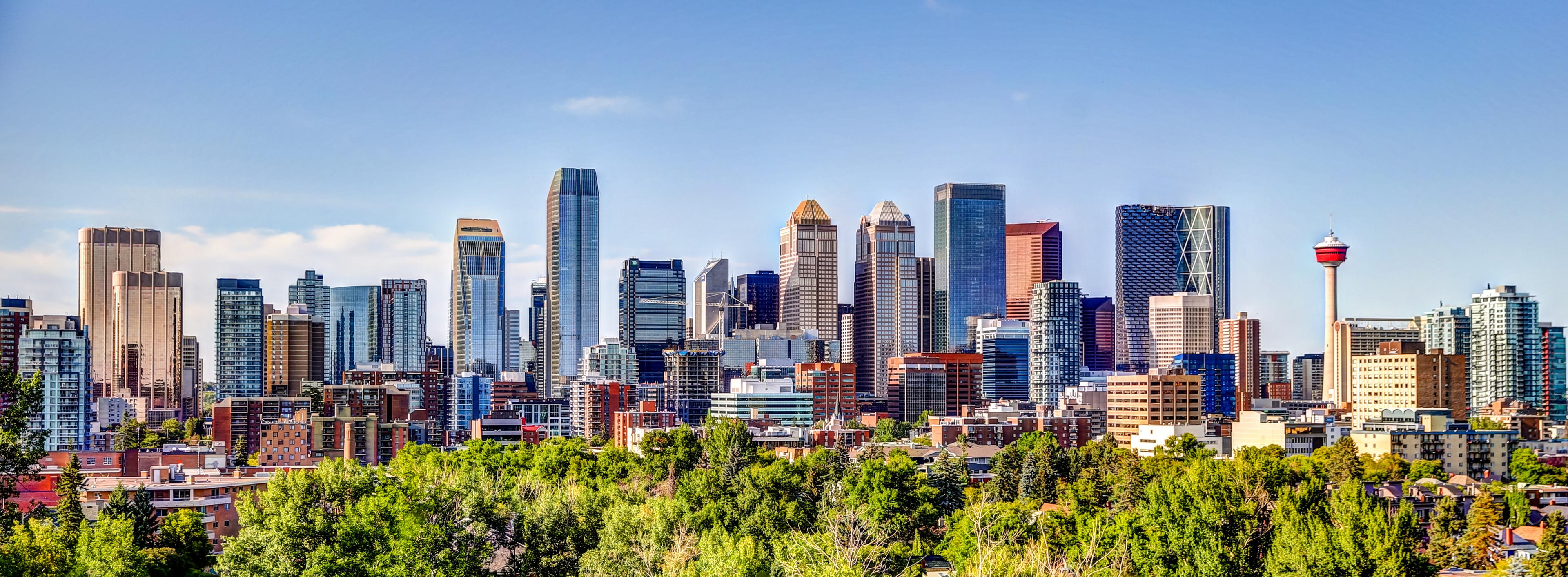 A vibrant view of Calgary's downtown skyline, featuring modern skyscrapers, the iconic Calgary Tower, and lush green trees in the foreground under a clear blue sky.