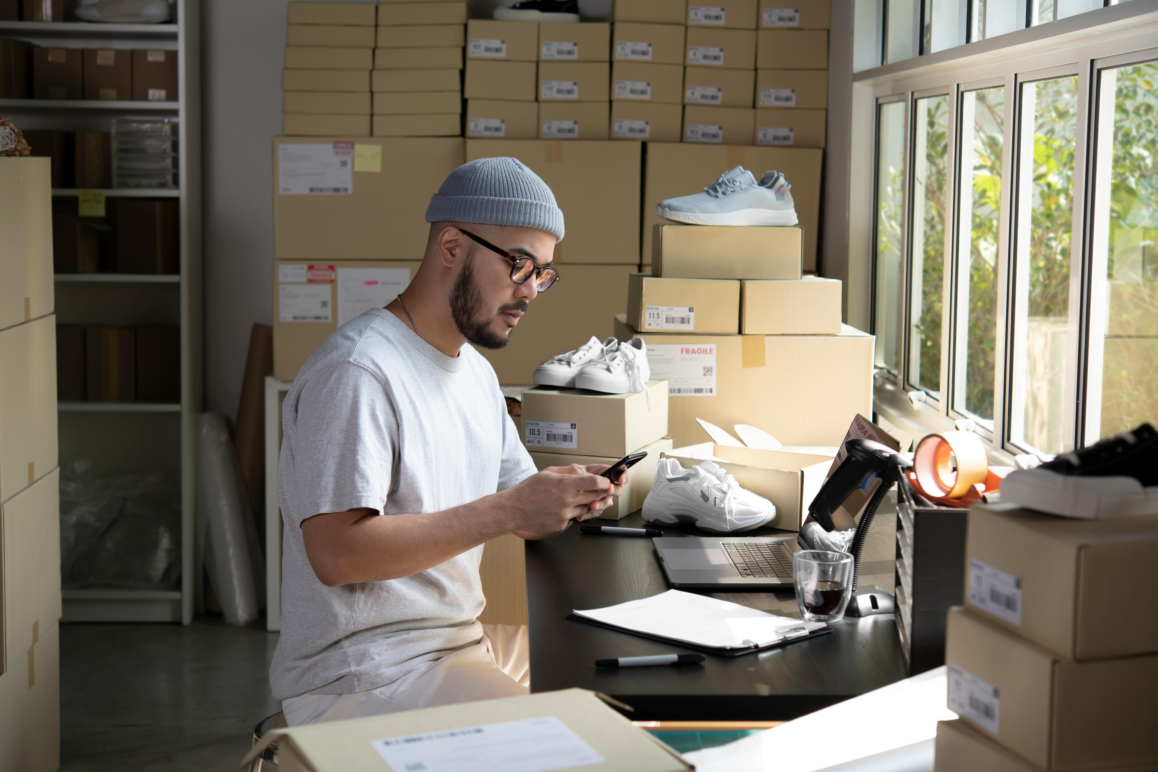 A man working in a room filled with shoe boxes, seated at a desk with a smartphone in hand. He is surrounded by packed and unpacked shoes, shipping materials, and a laptop, indicating a small business or e-commerce operation focused on footwear.