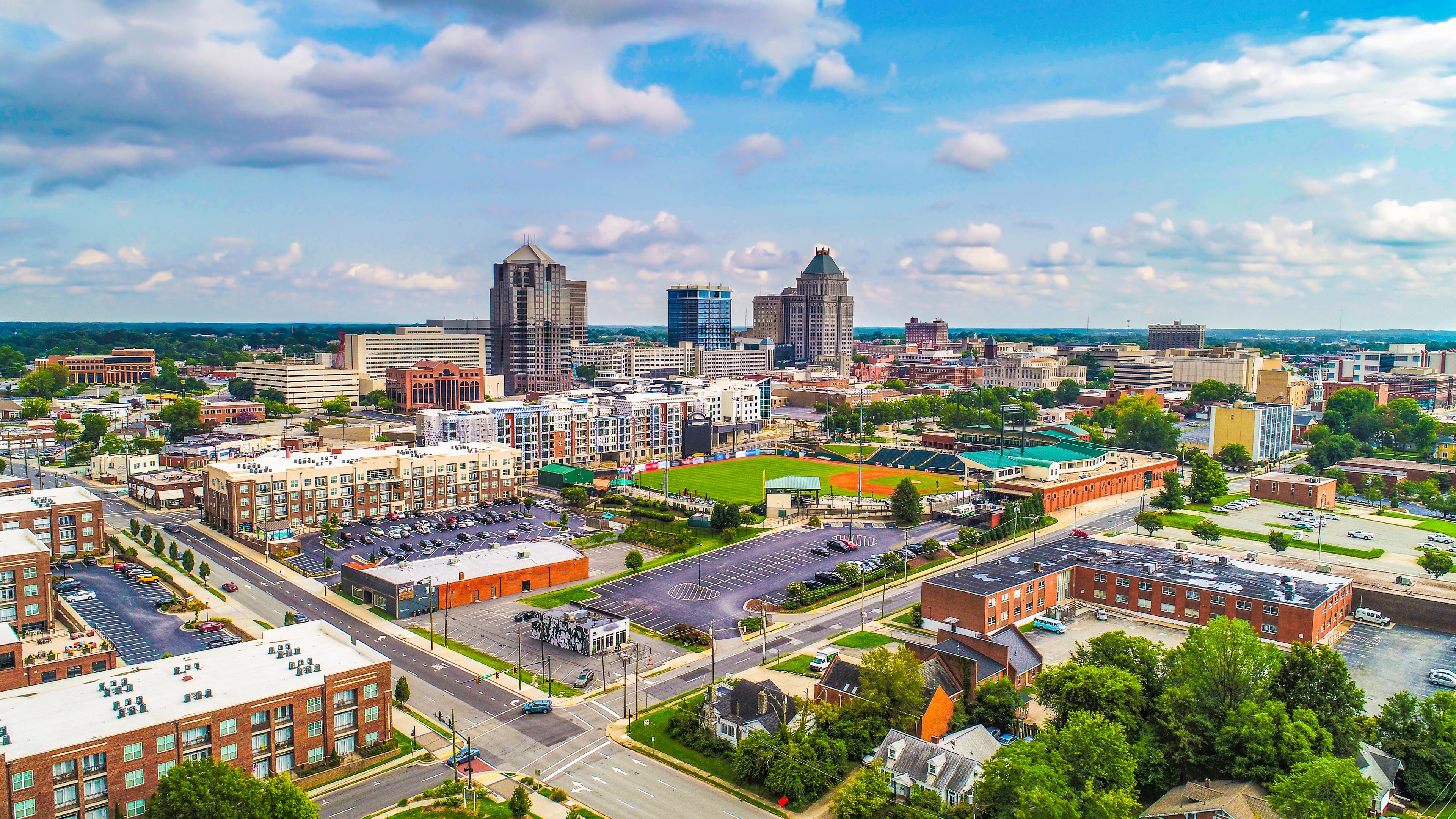 Panoramic aerial view of downtown Greensboro, North Carolina, featuring high-rise buildings, residential developments, and First National Bank Field baseball stadium under a partly cloudy sky.