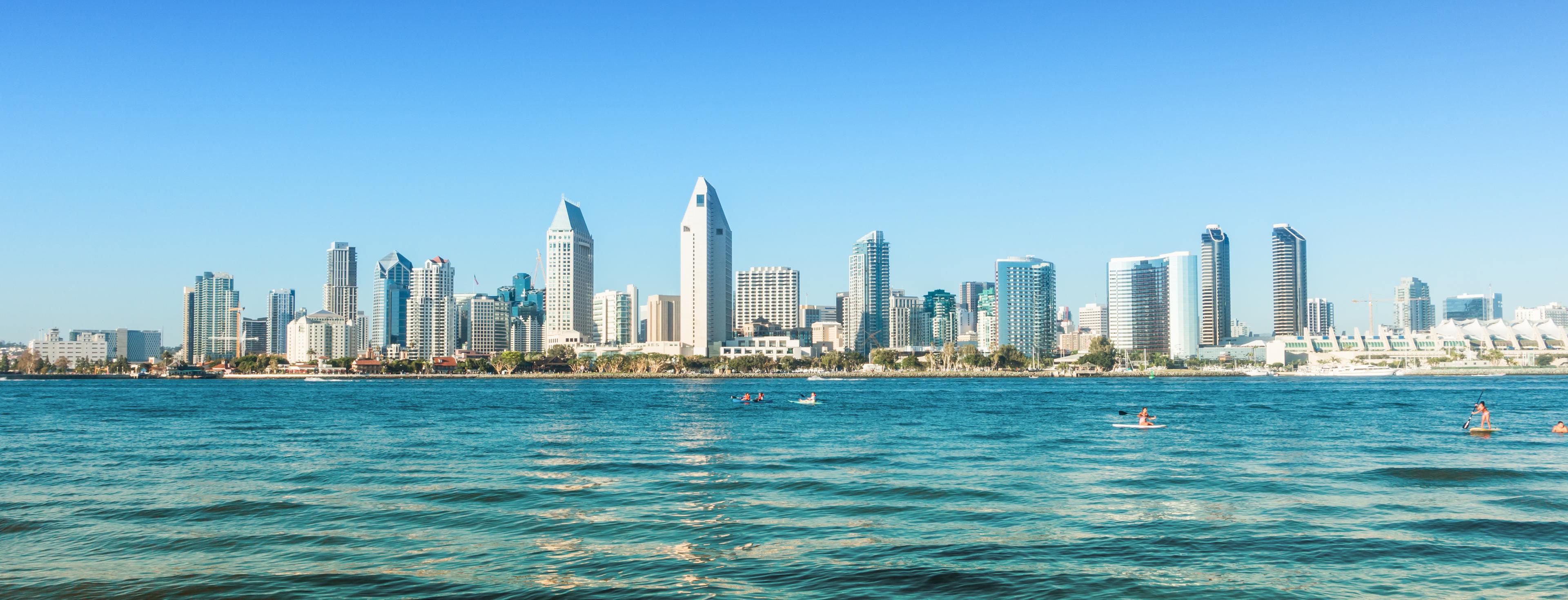Panoramic view of the San Diego skyline with clear blue skies and people enjoying water activities in the foreground.