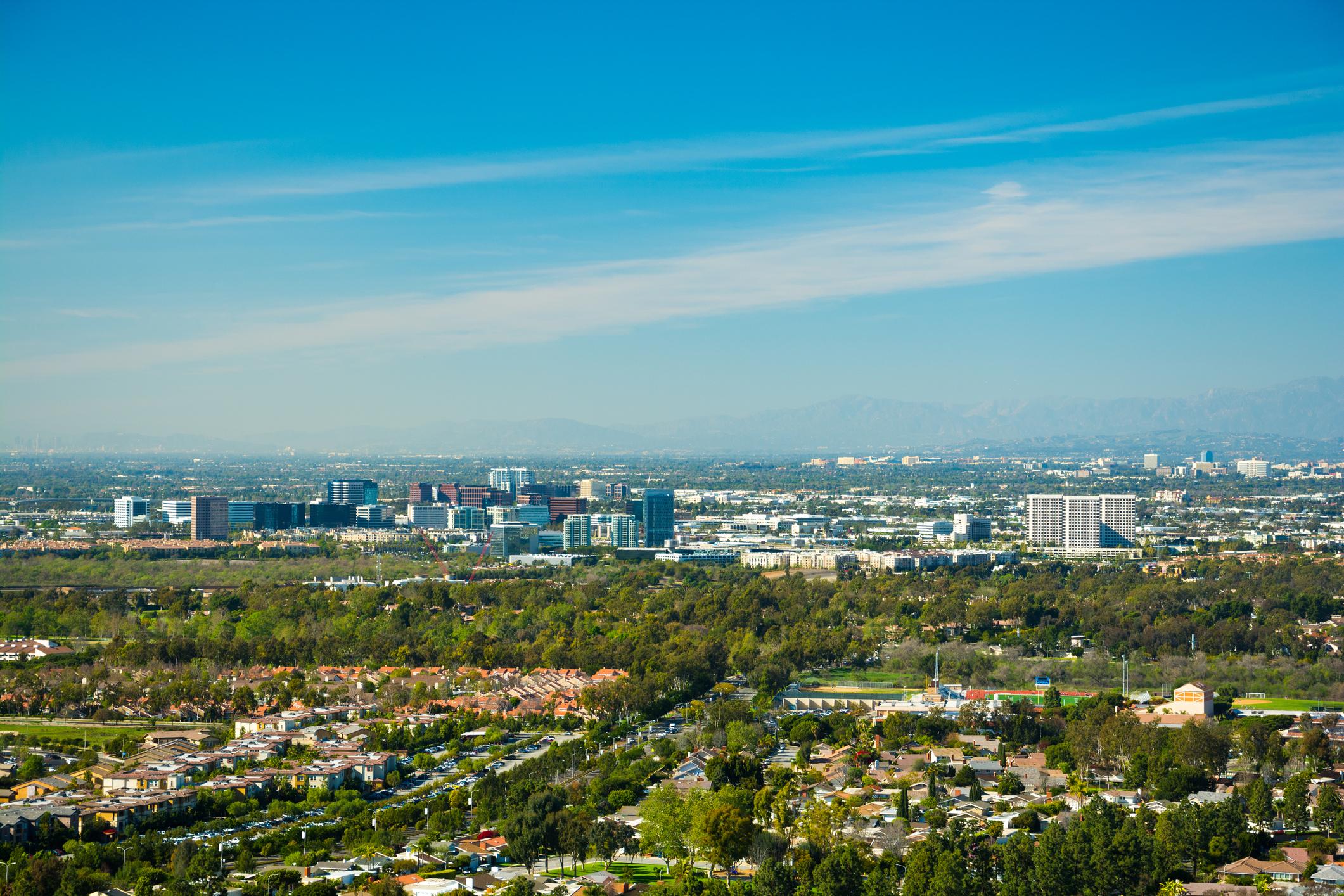 Elevated view of Irvine, California, highlighting suburban communities, corporate office buildings, and scenic mountain backdrops under a clear blue sky.