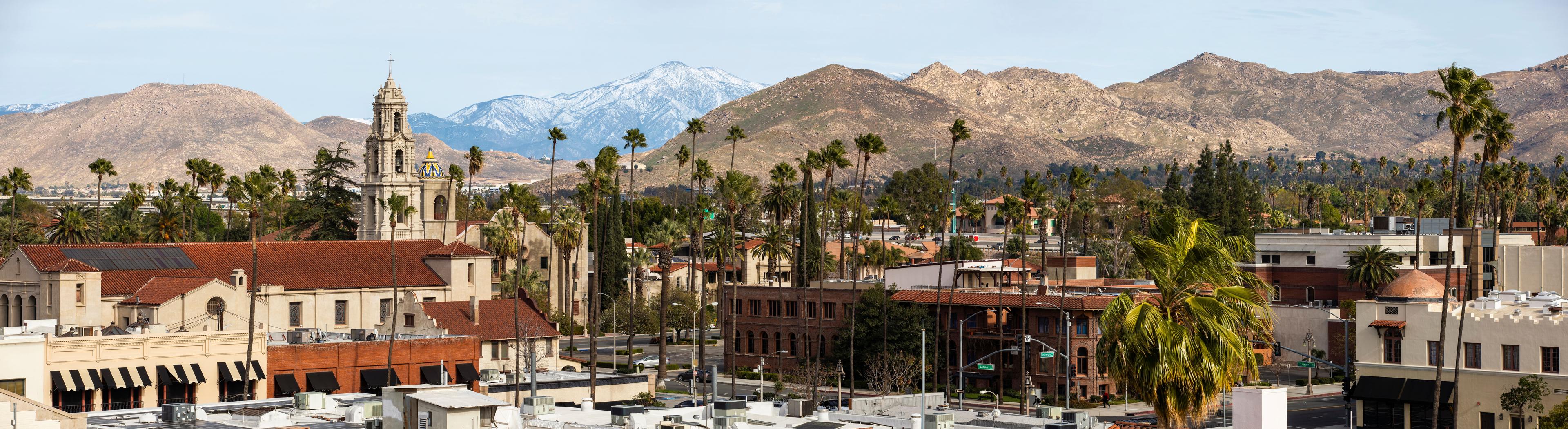 Riverside, CA cityscape framed by historic architecture, palm trees, and snowcapped mountains in the distance.