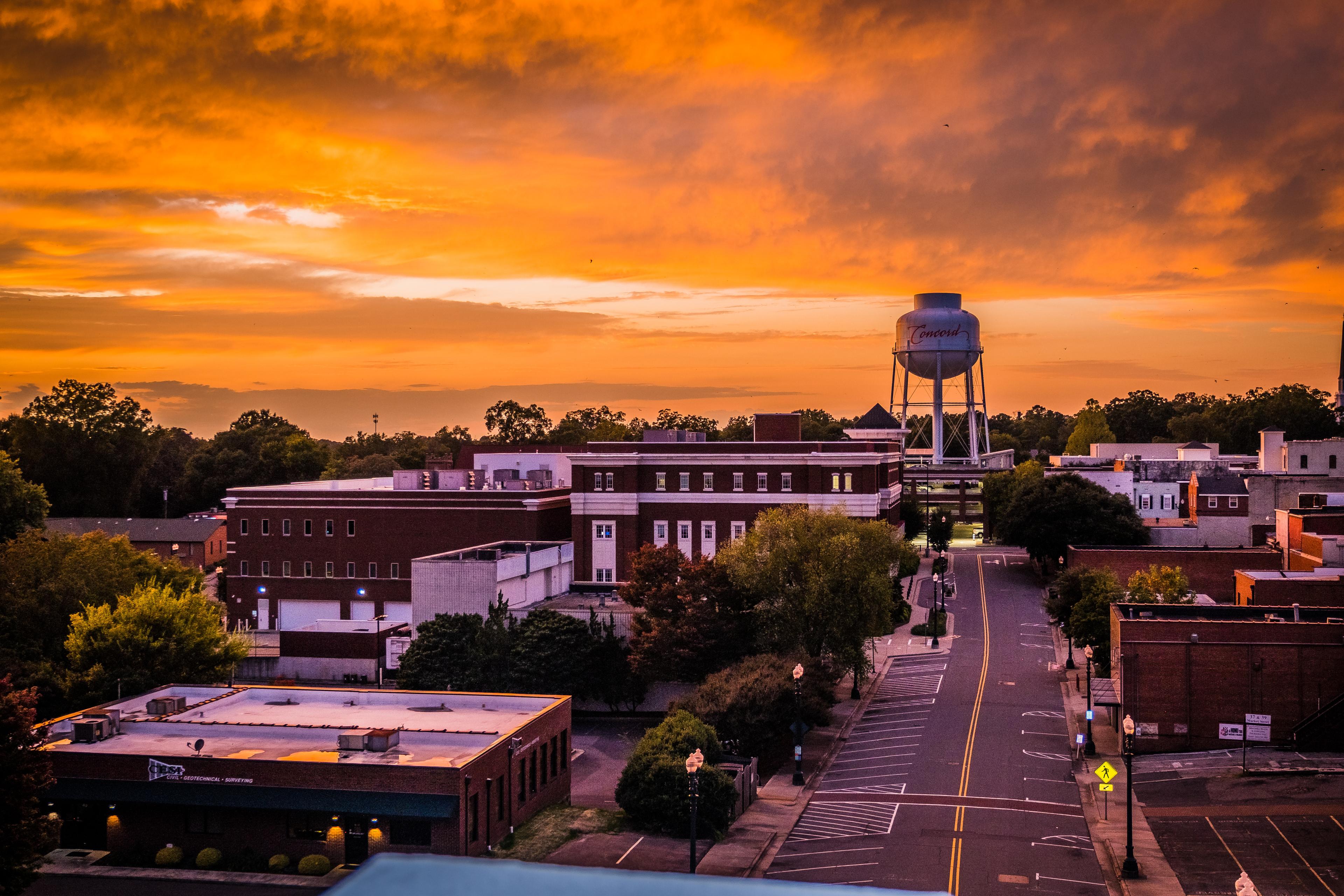 Vibrant sunset sky over downtown Concord, North Carolina, with the iconic water tower, tree-lined streets, and historic architecture.

