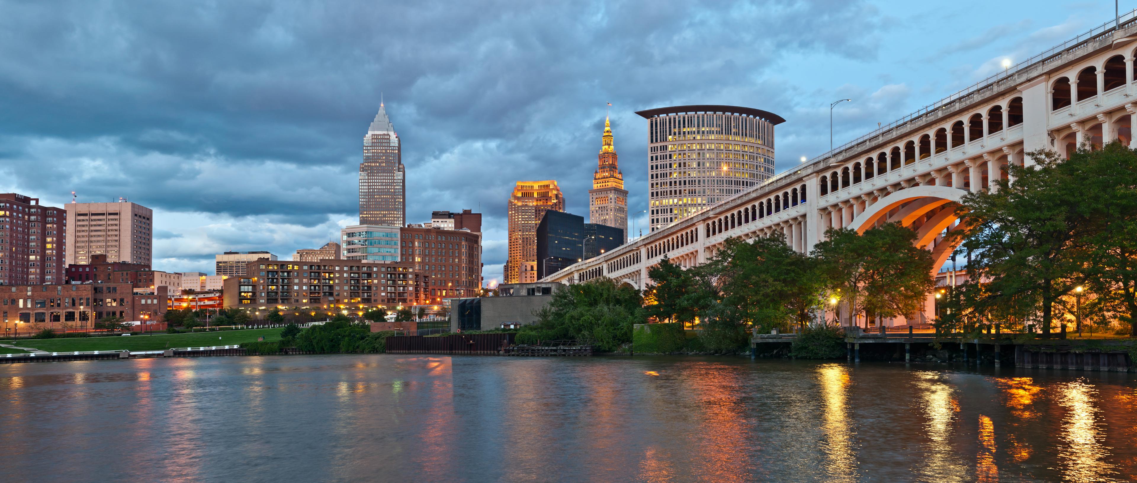 A twilight view of downtown Cleveland, Ohio, featuring a mix of historic and modern buildings, a brightly lit arched bridge, and the reflective waters of the Cuyahoga River under a cloudy sky.