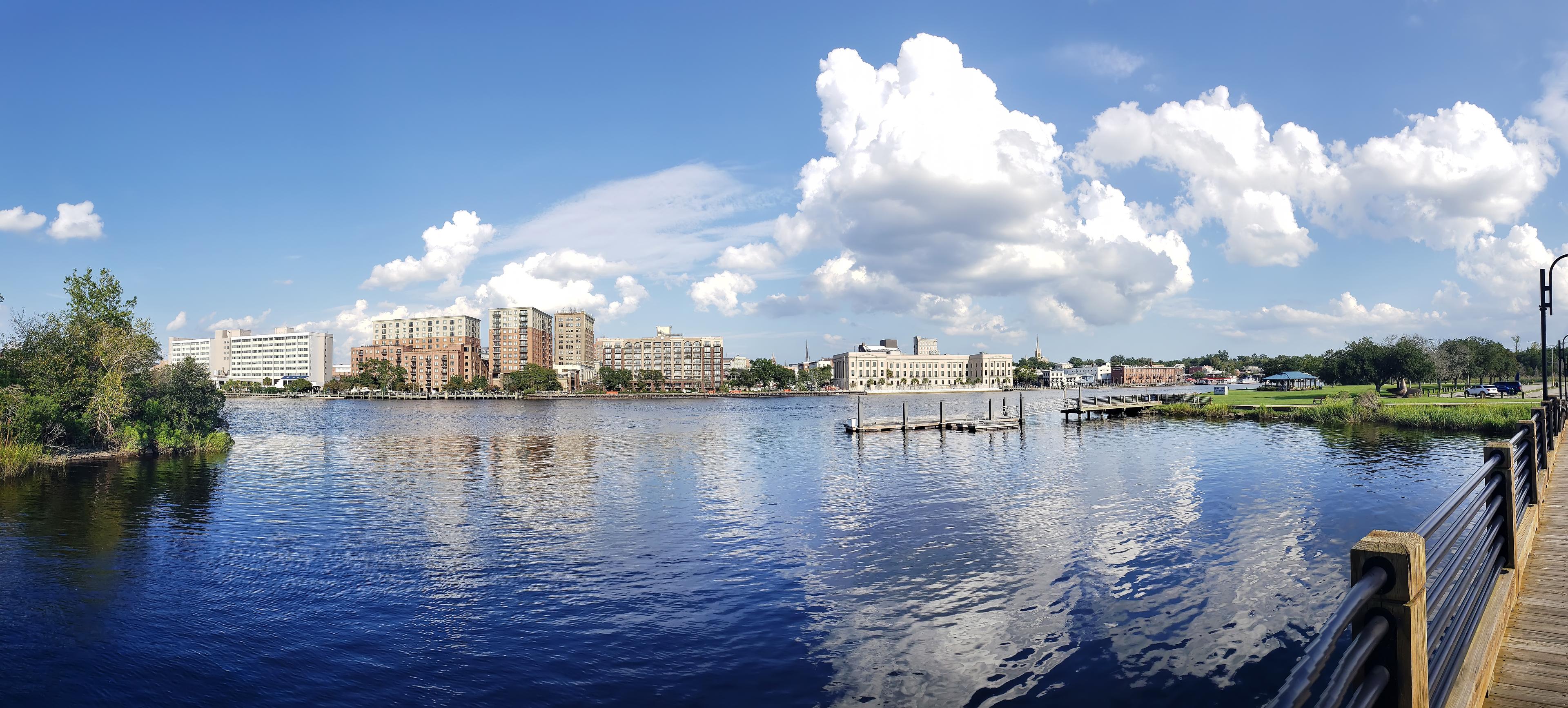 Panoramic view of Wilmington, North Carolina’s waterfront, featuring the Cape Fear River, downtown skyline, and historic architecture under a bright blue sky.