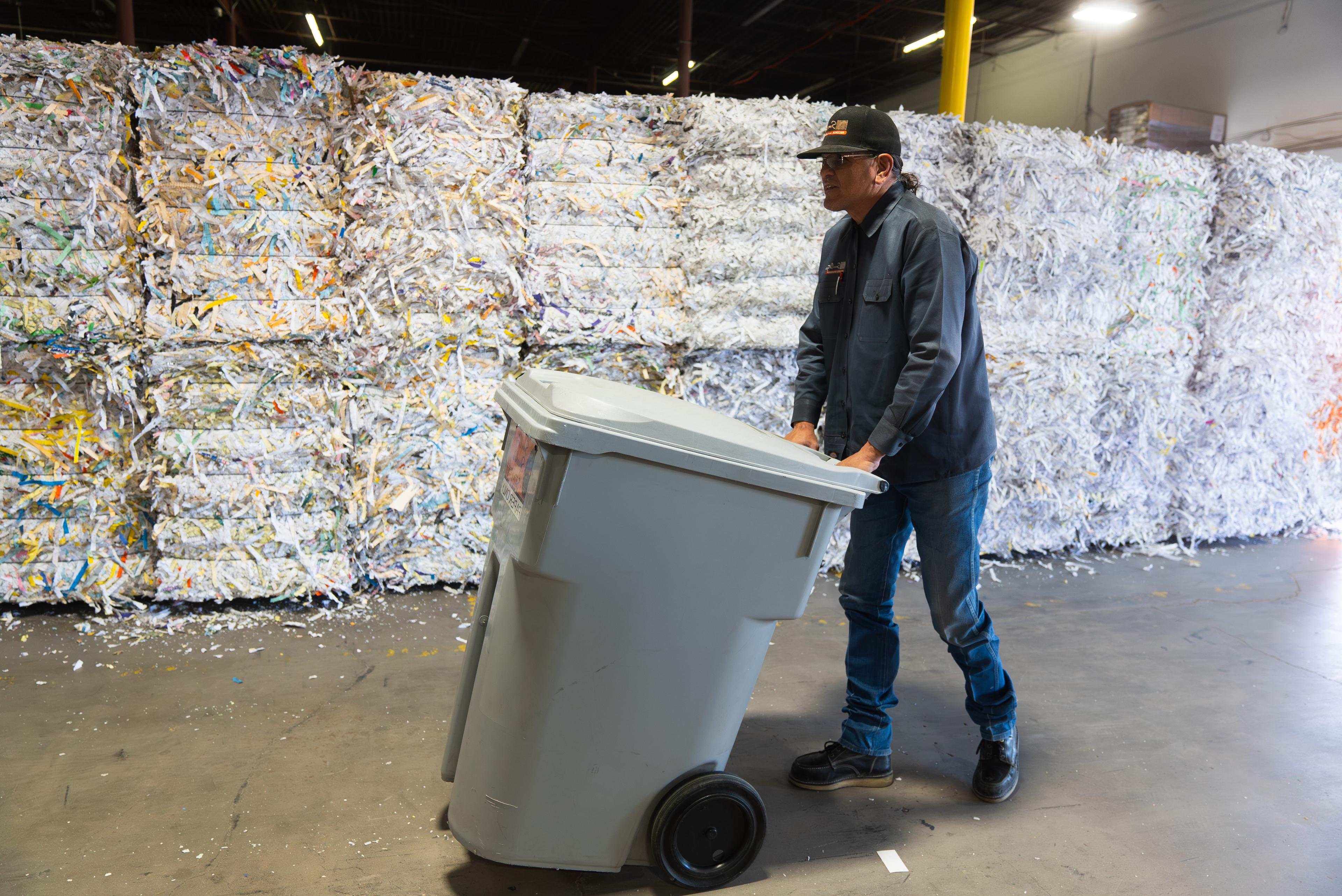 Worker transports a secure shred bin through a facility filled with baled shredded paper, showcasing industrial-scale document destruction and recycling operations.

