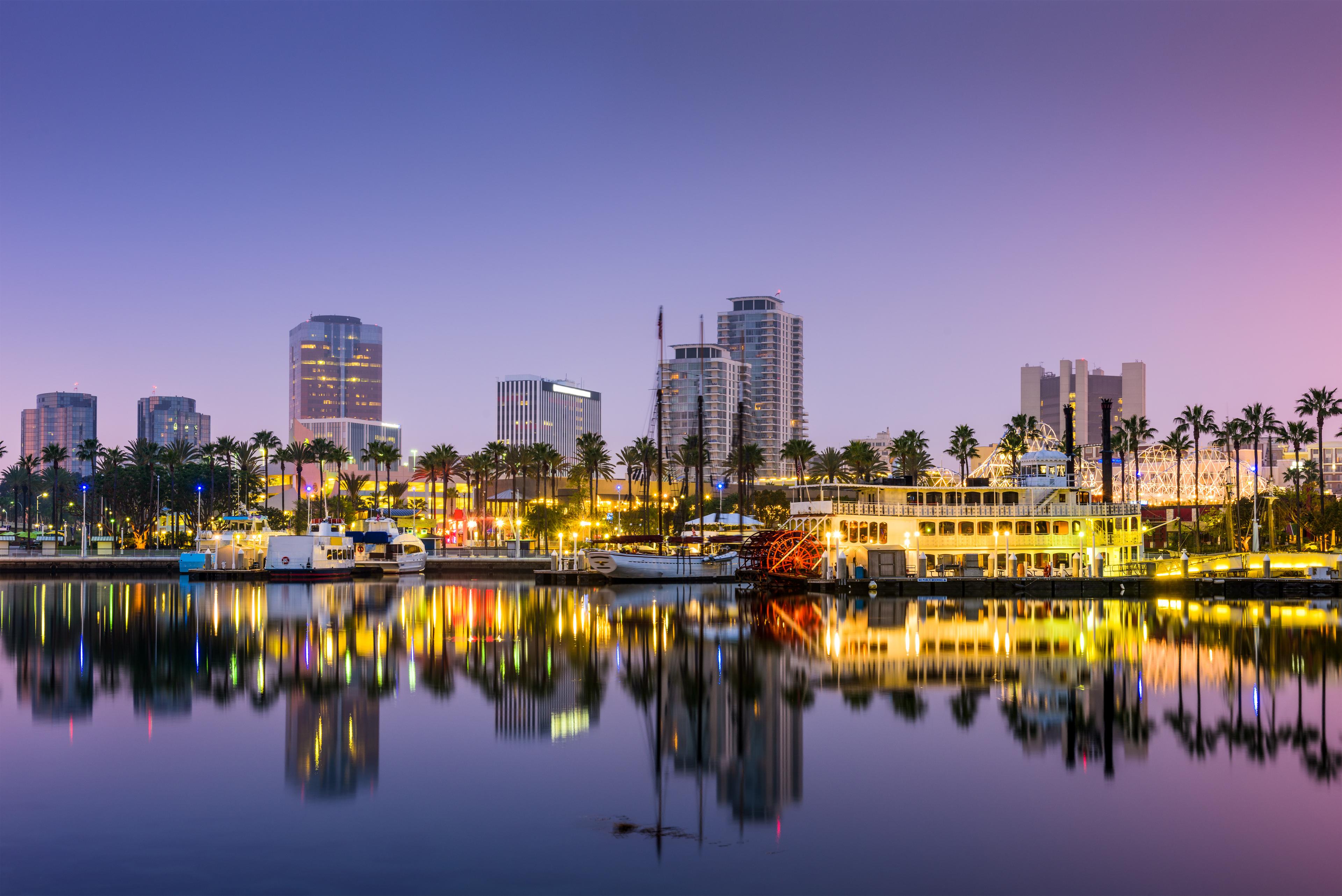 Evening view of the Long Beach, California waterfront featuring illuminated skyline, palm trees, harbor boats, and city lights reflecting off the calm water.