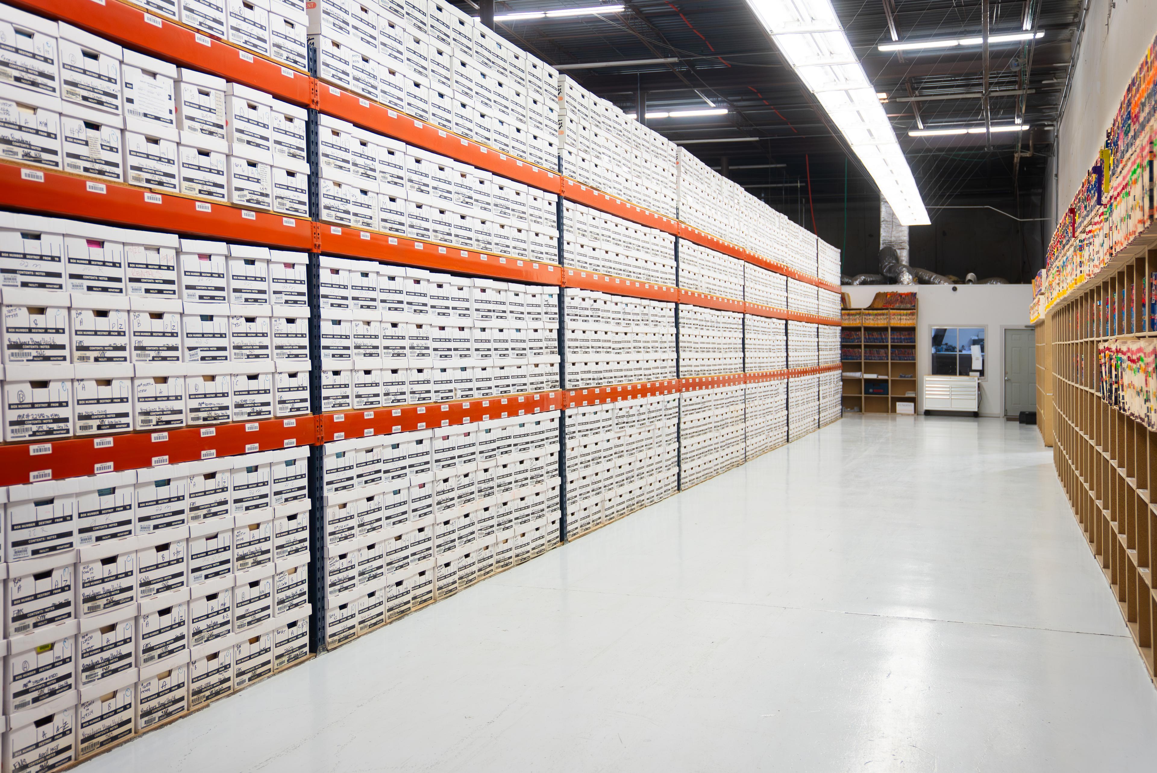 A record center aisle filled with shelves of neatly stacked archive boxes on one side, and shelves of file folders on the other side.


